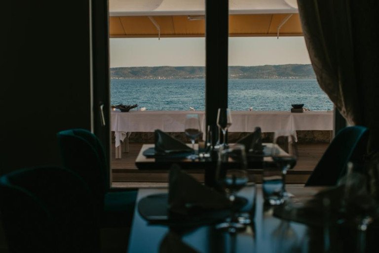Elegantno postavljen stol s čašama za vino u unutrašnjosti restorana Mala Mora, s pogledom kroz prozor na terasu tik uz plavo more i dalmatinsku obalu u pozadini