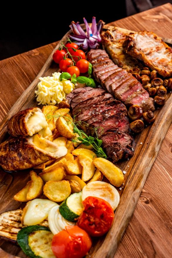 Bogata i masivna mesna plata na velikom drvenom pladnju s narezanim steakovima, roštiljem, pečenim krumpirima, šampinjonima i grilanim povrćem