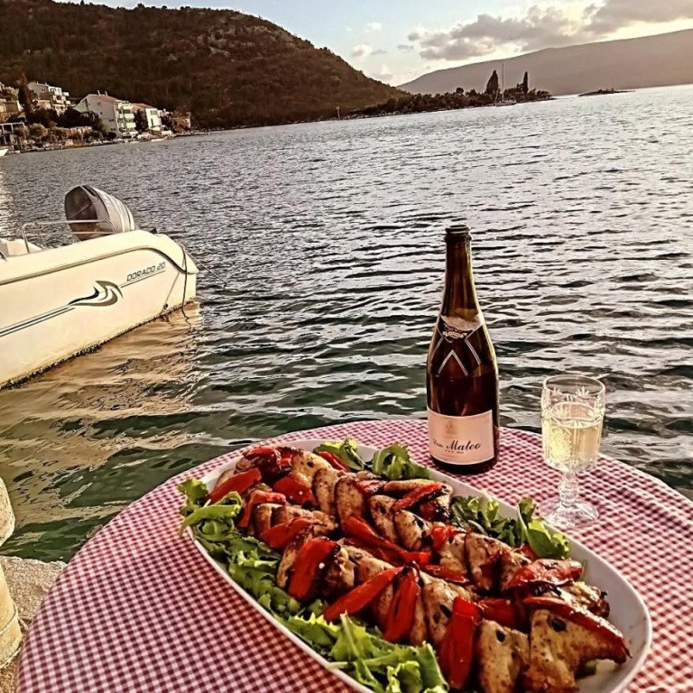Plata s mesom na žaru i povrćem na stolu s kariranim stolnjakom tik uz more, uz bocu i čašu bijelog vina te brodicu vezanu uz rivu